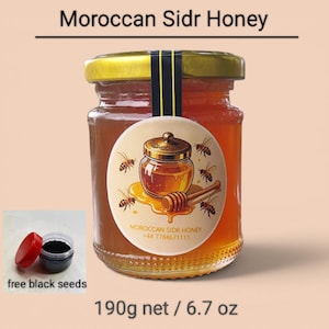 Könnte beinhalten: Ein Glasgefäß mit marokkanischem Sidr-Honig mit goldenem Deckel und einem Etikett mit einer Honigtopf-Illustration, Bienen und dem Text Moroccan Sidr Honey +44 7784671111. Daneben steht ein kleiner Behälter mit schwarzen Samen mit einem roten Deckel.
