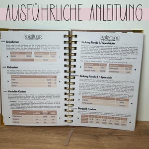 May include: Open notebook with gold spiral binding, featuring detailed financial planning pages. The pages include sections for income, fixed costs, and variable costs, with the title "AUSF&Uuml;HRLICHE ANLEITUNG" at the top.