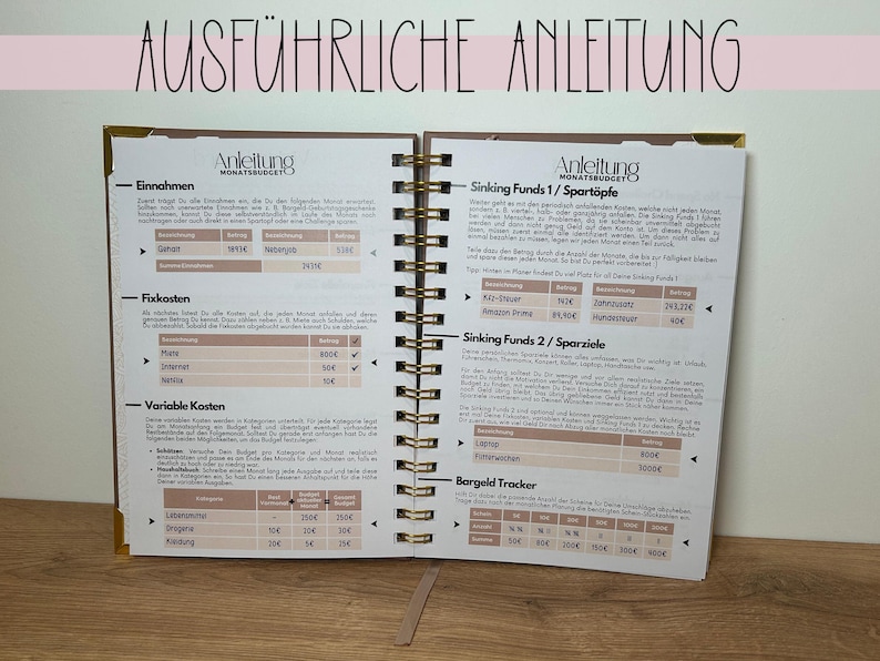 May include: Open notebook with gold spiral binding, featuring detailed financial planning pages. The pages include sections for income, fixed costs, and variable costs, with the title "AUSF&Uuml;HRLICHE ANLEITUNG" at the top.