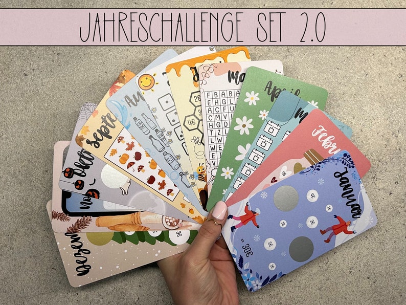 May include: A set of 12 colourful cards with different designs for a yearly challenge. The cards feature various themes, including a beehive, a snowman, a pumpkin, and a bottle of wine. The text on the cards reads "JAHRESCHALLENGE SET 2.0", "Mo", "Feb", "Januar", "Dezen", "Septe", "Ay", "30&euro;", and "26".
