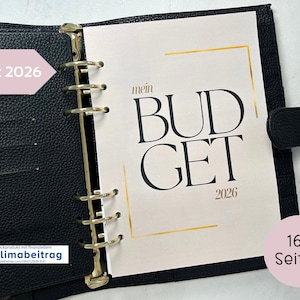 Peut inclure: Un agenda en cuir noir ouvert sur une page avec "BUDGET 2026" en grandes lettres noires. La page a des accents dorés et est maintenue par des anneaux dorés. L'agenda a également une fermeture à sangle noire.