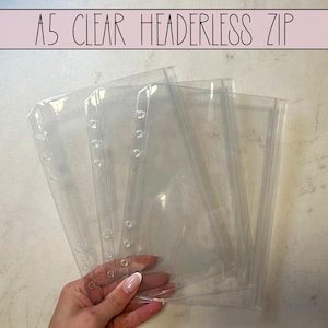 Könnte beinhalten: Transparente A5-Reißverschlusstaschen ohne Kopf. Die transparenten Kunststofftaschen haben vorgestanzte Löcher für Binderinge. Der Text "A5 CLEAR HEADLERLESS ZIP" ist in Schwarz auf rosa Hintergrund gedruckt.