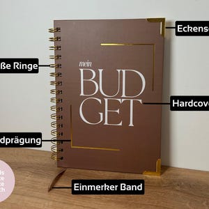 May include: A brown spiral-bound notebook with the word "BUDGET" in white lettering. The notebook has gold corner protectors and a gold-colored spiral binding. The cover also features gold accents and the text "mein".