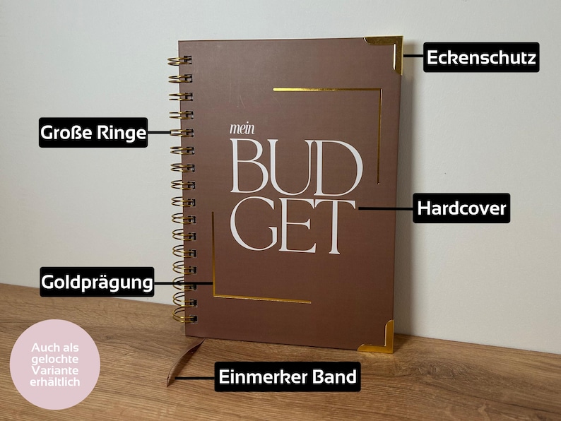May include: A brown spiral-bound notebook with the word "BUDGET" in white lettering. The notebook has gold corner protectors and a gold-colored spiral binding. The cover also features gold accents and the text "mein".