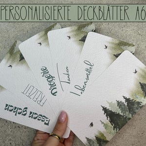 Op de afbeelding: Vijf gepersonaliseerde A6-kaarthouders met een aquarel-bosontwerp. De tekst op de houders luidt "Essen & Trinken", "Freizeit", "Drogerie", "Tanken" en "Lebensmittel".