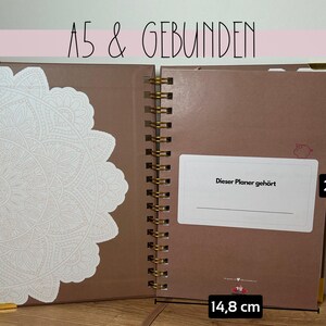 May include: Open A5 size planner with a brown cover and gold spiral binding. The planner is 21 cm tall and 14.8 cm wide. The left side has a white decorative design. The text "Dieser Planer geh&ouml;rt" is visible.