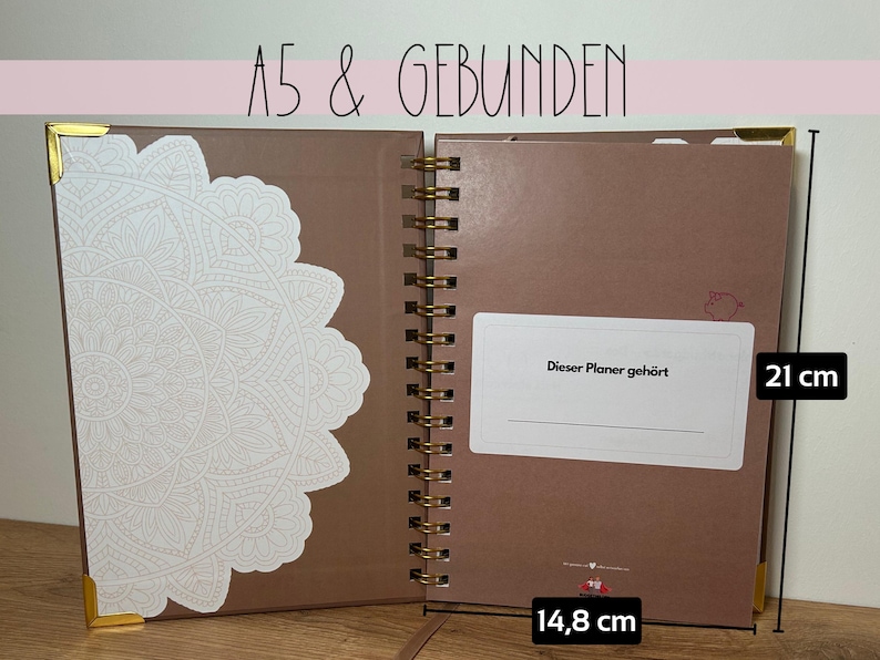 May include: Open A5 size planner with a brown cover and gold spiral binding. The planner is 21 cm tall and 14.8 cm wide. The left side has a white decorative design. The text "Dieser Planer geh&ouml;rt" is visible.