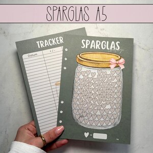 Könnte beinhalten: Ein grauer und weißer A5-Tracker mit einem grauen und weißen Glas mit Herzen darauf. Der Tracker hat das Wort "Tracker" und die Wörter "Datum" und "+/-" darauf gedruckt. Das Glas hat das Wort "Sparglas" darauf gedruckt.