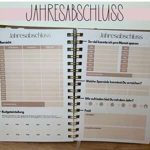 May include: Open notebook with the German text "JAHRESABSCHLUSS" at the top. The pages are filled with tables and prompts for financial planning, including sections for income, expenses, and savings goals. The notebook has a spiral binding.