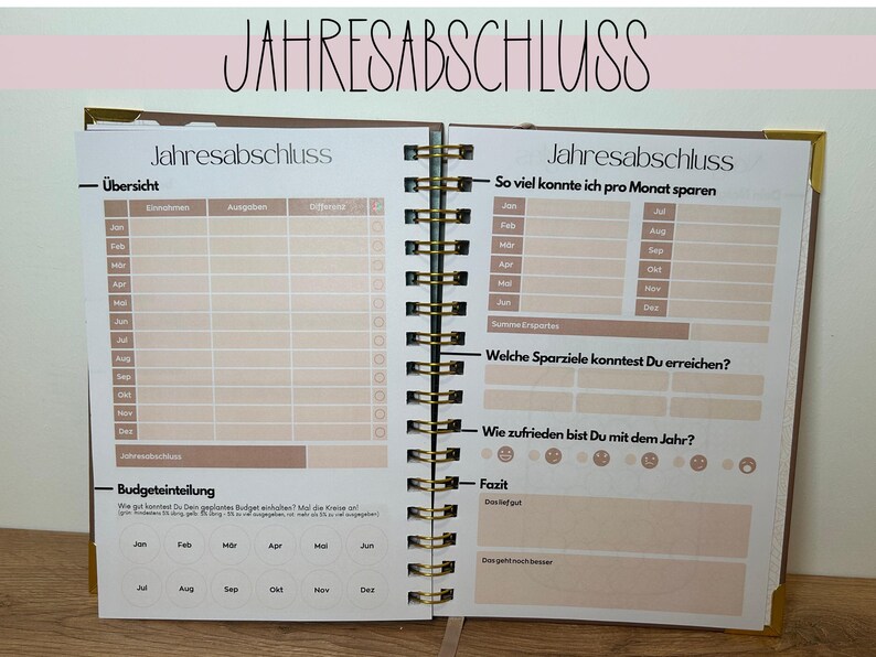 May include: Open notebook with the German text "JAHRESABSCHLUSS" at the top. The pages are filled with tables and prompts for financial planning, including sections for income, expenses, and savings goals. The notebook has a spiral binding.
