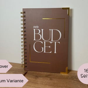 May include: A brown hardcover budget planner with gold accents and spiral binding. The cover features the words "mein BUDGET" in white. The planner has 160 pages.