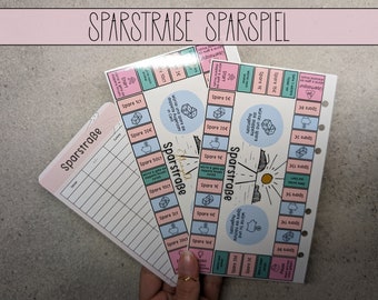 Pode incluir: Dois tabuleiros de jogo coloridos com o título "Sparstraße Sparspiel" e uma folha de rastreamento de poupança rosa e branca. Os tabuleiros de jogo apresentam um tema de poupança com ilustrações de cofrinhos, moedas e dados.
