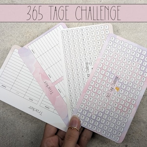Könnte beinhalten: Eine Hand hält mehrere weiße Tracker-Karten mit einem rosa-lila Aquarelldesign. Die Karten haben ein Rasterlayout zur Fortschrittsverfolgung, mit dem Text "365 Tage Challenge" oben. Das Wort "Tracker" ist ebenfalls sichtbar.