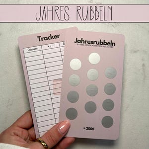 Könnte beinhalten: Zwei rosa Karten mit dem Text "JAHRES RUBBELN" oben. Eine Karte ist ein Tracker mit Linien und dem Wort "Tracker". Die andere Karte hat Rubbelkreise und den Text "Jahresrubbeln". Die Karten werden von einer Person gehalten.