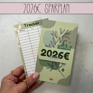 May include: A green and white savings tracker with the text "2026€ Sparplan" and "Tracker". The tracker has a floral design and a space to write the date and amount. The tracker is held by a person.
