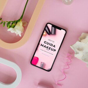 Può includere: Uno smartphone che mostra un download digitale rosa e bianco per un piano di trucco chiamato "Guida Makeup". Il testo sullo schermo recita "Makeup Plan Guida Makeup 8 Piano Makeup di 8 Settimane www.makeupdaily.com".