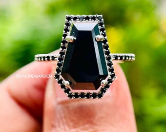 May include: A silver ring featuring a large, black, coffin-shaped gemstone. The gemstone is surrounded by a border of small, black stones. The ring band also has small black stones. The ring is held up against a blurred green background.