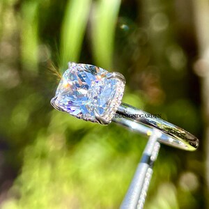 May include: A silver ring with a large, clear, cushion-cut diamond. The diamond is set in a pave setting with smaller diamonds around the edges. The ring is on a white background.