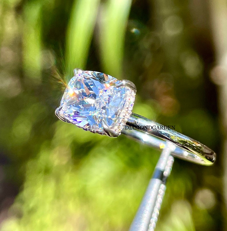 May include: A silver ring with a large, clear, cushion-cut diamond. The diamond is set in a pave setting with smaller diamonds around the edges. The ring is on a white background.