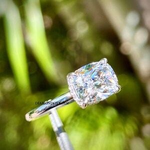 May include: A cushion-cut diamond engagement ring set in a white gold band with a halo of smaller diamonds. The ring is on a display stand against a green background.