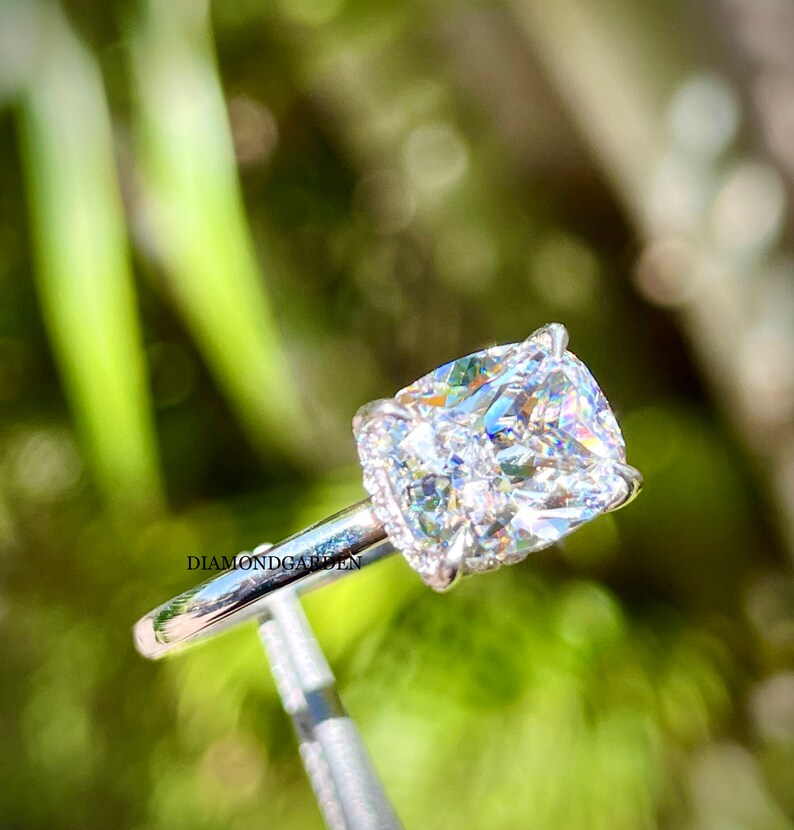 May include: A cushion-cut diamond engagement ring set in a white gold band with a halo of smaller diamonds. The ring is on a display stand against a green background.