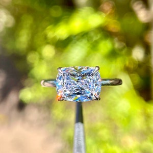 May include: A cushion-cut diamond engagement ring set in a silver band. The diamond is clear and sparkly, and the ring is simple and elegant.