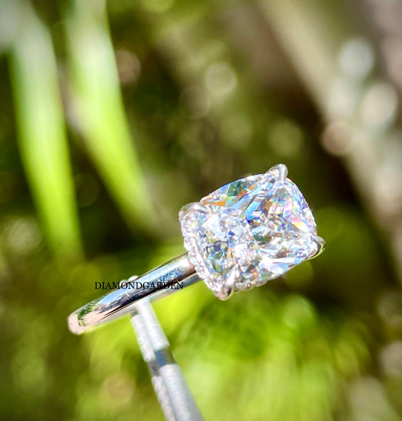 May include: A cushion-cut diamond engagement ring set in a white gold band with a halo of smaller diamonds. The ring is on a display stand against a green background.