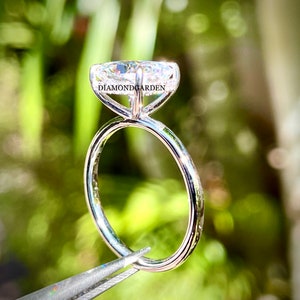 May include: A silver solitaire engagement ring with a large, clear, oval-shaped diamond. The ring is held by tweezers and the text "DIAMONDGARDEN" is visible on the band.