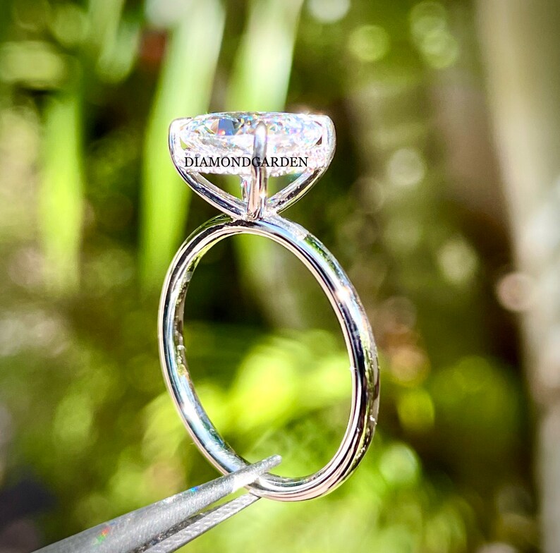 May include: A silver solitaire engagement ring with a large, clear, oval-shaped diamond. The ring is held by tweezers and the text "DIAMONDGARDEN" is visible on the band.
