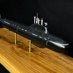 May include: A detailed black model submarine with white accents, mounted on brass supports and a wooden base. The model features intricate detailing, including a propeller, periscopes, and other navigational elements. The submarine is set against a dark background.