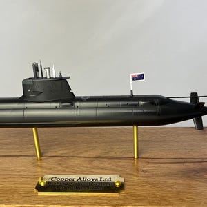 Puede incluir: Maqueta de submarino negro con una pequeña bandera australiana, montada sobre soportes dorados. El modelo se exhibe sobre una base de madera con una placa dorada que dice "Copper Alloys Ltd".
