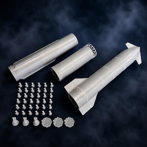 May include: A model rocket kit, featuring various components in light grey. The kit includes cylindrical sections, a nose cone, fins, and numerous small engine nozzles. The parts are arranged on a dark blue background.