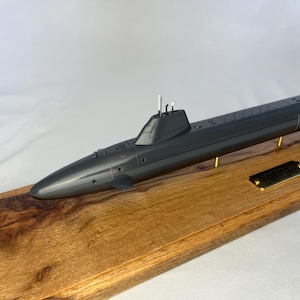 May include: A dark gray model of a submarine on a wooden base with a brass plaque. The submarine has a periscope and a small flag on the back.