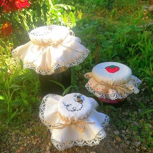 Peut inclure: Trois pots de confiture en verre, chacun avec un couvercle en tissu blanc, attachés avec de la ficelle. Un couvercle présente une fraise, un autre une abeille et le troisième un motif cousu. Les pots sont posés sur un lit de cailloux et d'herbe, avec des fleurs rouges en arrière-plan.