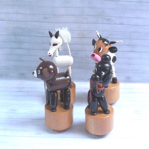 May include: Four small wooden animal figurines standing on cylindrical bases. The set includes a white horse with a white mane, a brown dog, a black poodle with a pink collar, and a black and white cow with a pink nose and small horns.