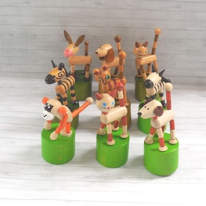 Op de afbeelding: Een verzameling houten dierenspeelgoed, waaronder een ezel, een hond, een kat, een zebra en een aap. Elk speeltje is beschilderd met heldere kleuren en gemonteerd op een groene basis. De speeltjes hebben beweegbare ledematen.