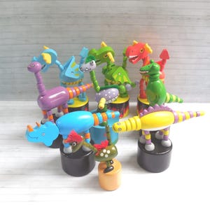 May include: A collection of colorful wooden push-up toys featuring various dinosaur and dragon figures. Each toy stands on a cylindrical base and displays vibrant hues including purple, blue, green, yellow, and red with painted details.