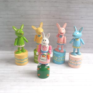 May include: A collection of five colorful wooden bunny figurines. Each bunny is painted in a different pastel color, including green, yellow, pink, and blue. The bunnies stand on cylindrical bases, decorated with painted patterns.