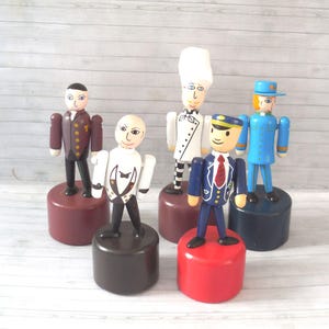 May include: A collection of six wooden toy figures depicting various professions. Each figure is painted with detailed clothing and facial features, standing on a cylindrical base. The figures include a butler, chef, and a pilot.