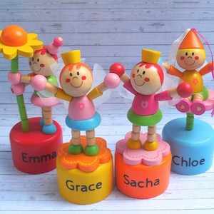 May include: Four colorful wooden puppet figures with different names on their bases: Emma, Grace, Sacha, and Chloe. Each figure has a different design and color scheme, with details like wings, flowers, and crowns.