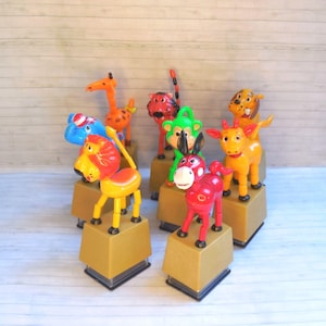 May include: A collection of seven colorful plastic animal figurines, each mounted on a square tan base. The set includes a red donkey, a yellow lion, a green monkey, a blue elephant, an orange giraffe, a tiger, and a brown dog with spots.