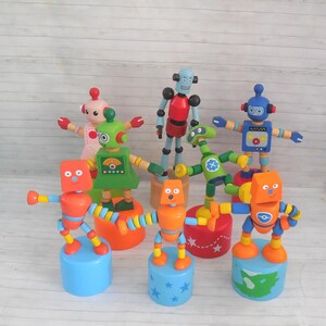 May include: A collection of seven colorful wooden robot toys, each mounted on a cylindrical base. The robots feature various designs, including red, green, orange, and blue bodies with painted details like gears, buttons, and stars on the bases.