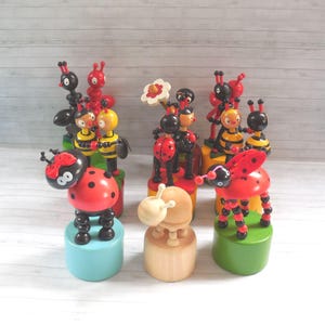 May include: A selection of brightly coloured wooden insect toys, including ladybirds, bees, and ants. The toys are painted in red, yellow, black, and blue, and are displayed on small, round bases. The toys are designed to be interactive.