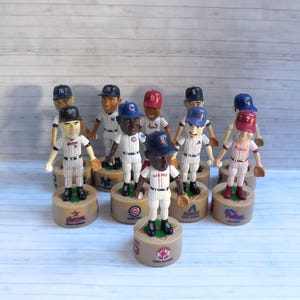May include: A collection of baseball bobblehead figures, each wearing a different team's uniform and cap. The figures stand on round bases with team logos and player names. The bobbleheads are in various colors, including red, blue, and white.