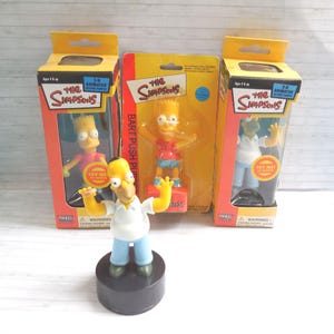 May include: A selection of Simpsons character figurines, including Homer, Bart, and others, in their original packaging. The figures are brightly coloured and feature the show's iconic characters. The packaging includes the text "The Simpsons".