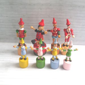 Peut inclure: Plusieurs jouets Pinocchio en bois. Les figurines portent des tenues rouges, jaunes, vertes et bleues, les bras tendus. Chaque jouet est posé sur une base cylindrique. Le fond est un grain de bois clair.