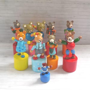 May include: A collection of nine wooden push-puppet toys featuring bear figures in various colours and outfits, each mounted on a cylindrical base. The bears have painted facial features and articulated limbs, displayed in a group on a light surface.