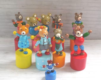 May include: A collection of nine wooden push-puppet toys featuring bear figures in various colours and outfits, each mounted on a cylindrical base. The bears have painted facial features and articulated limbs, displayed in a group on a light surface.