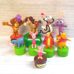 May include: A collection of colorful character figurines, including a lion, horse, Cat in the Hat, ant, Winnie the Pooh, Tigger, and Piglet. Each figure is mounted on a green or brown base. The Piglet figure is on a brown base with the name "Piglet".