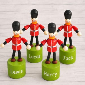 May include: Four wooden toy soldiers wearing red tunics, black bearskin hats, and black trousers, each mounted on a green cylindrical base. The bases are personalized with the white painted names Lewis, Lucas, Harry, and Jack in a script font.
